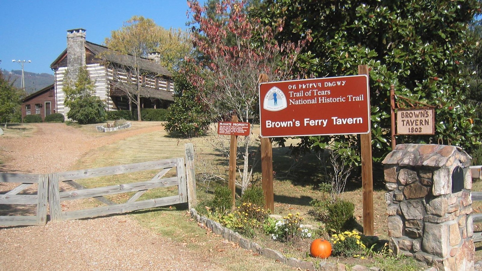 A brown sign next to a dirt road leading to a house in the distance.