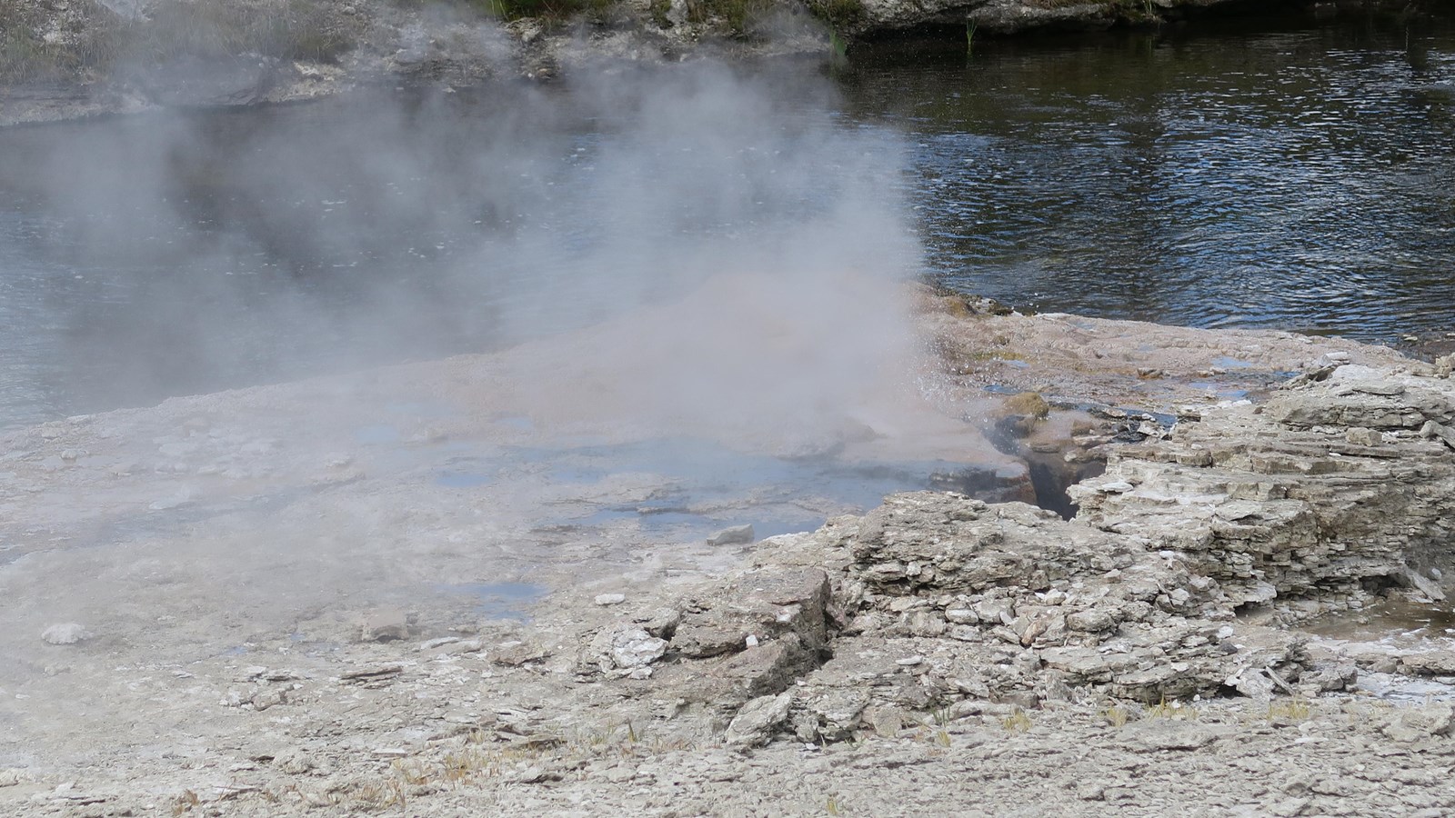 Steam rises from a tan shelf of rock in front of a river.