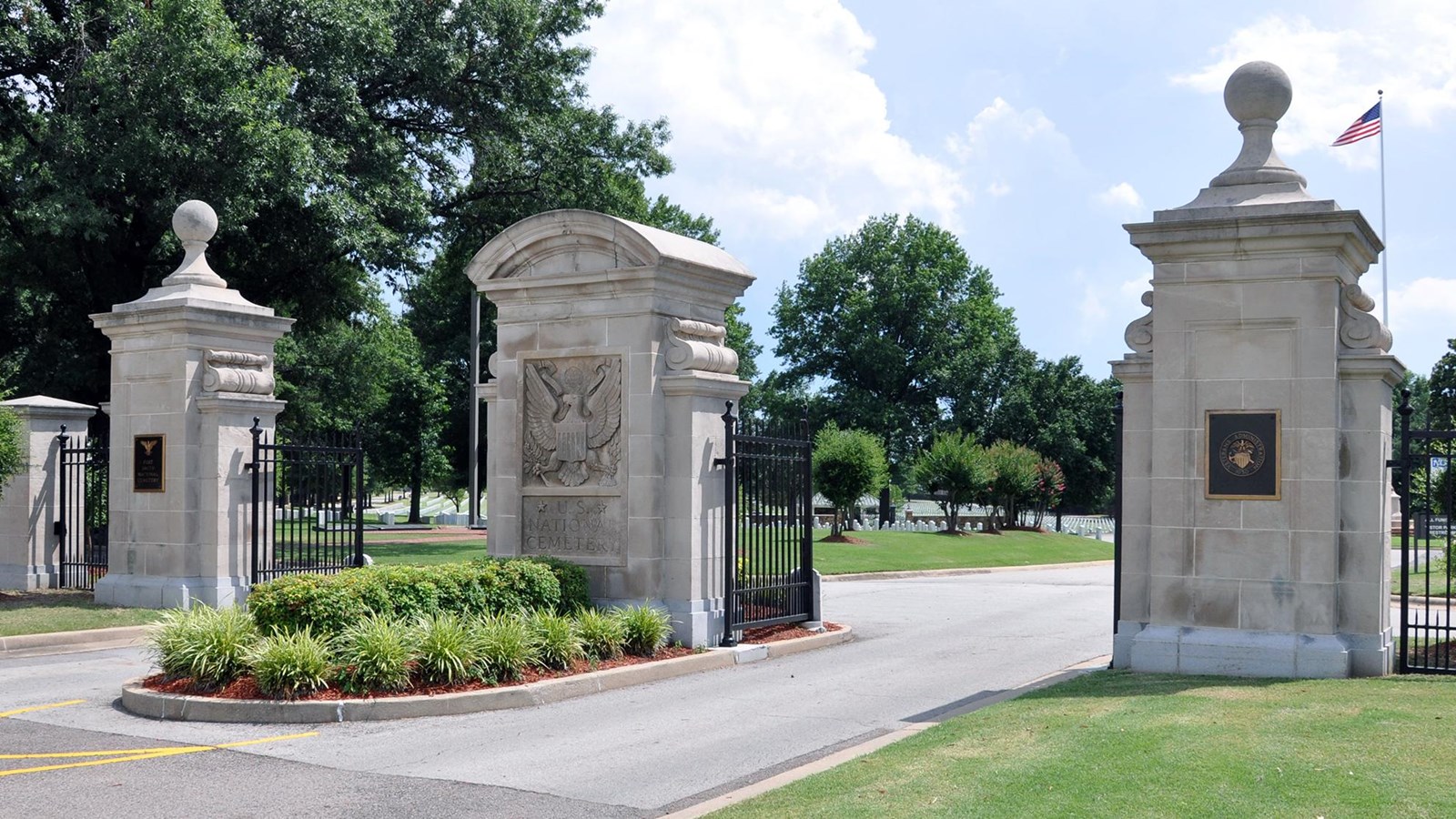 Three wide ornate gray stone ten foot square columns form a divided drive entrance to the cemetery. 