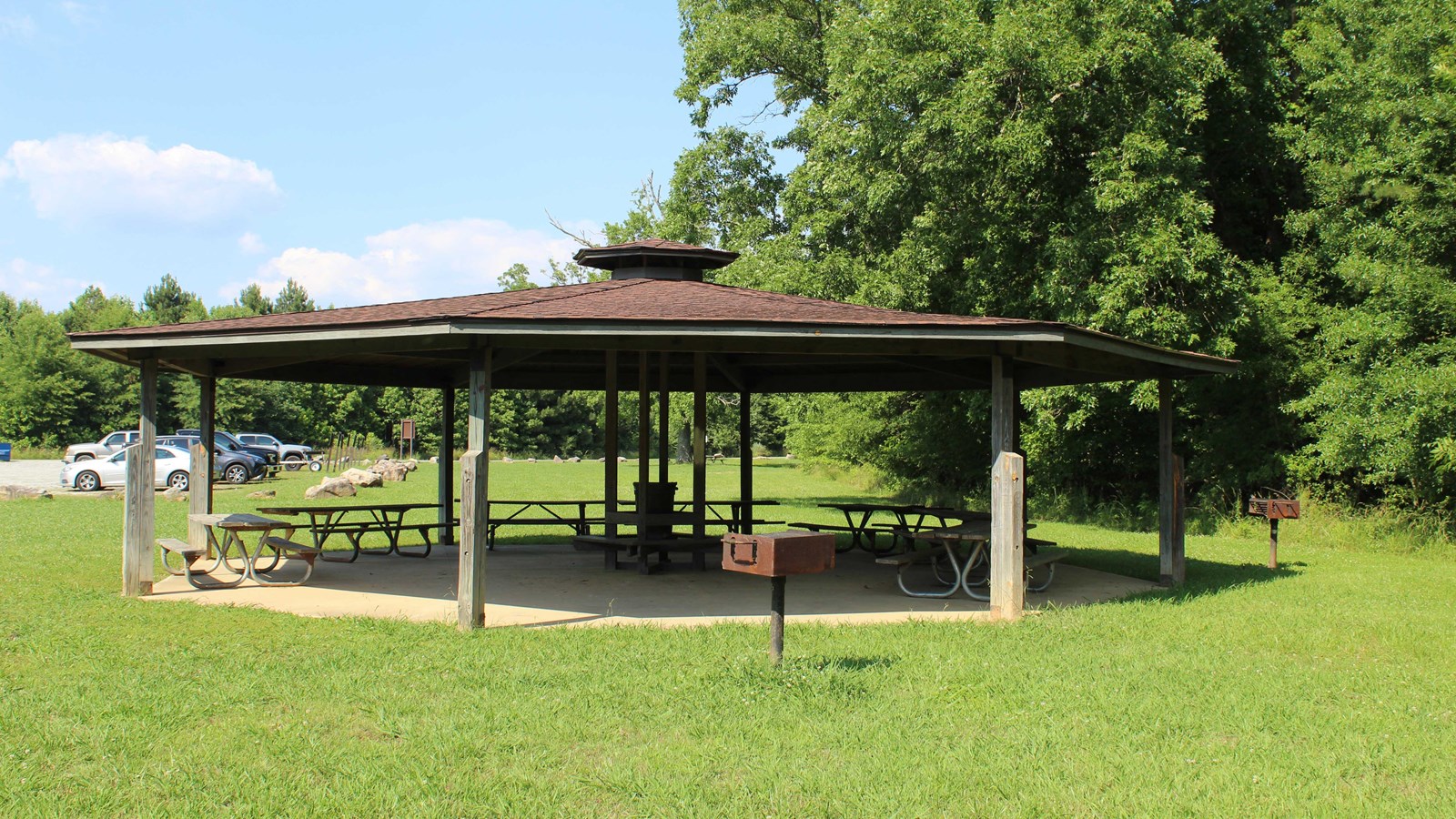 Octagonal shaped open air pavilion with picnic tables and grills with parking area in background.