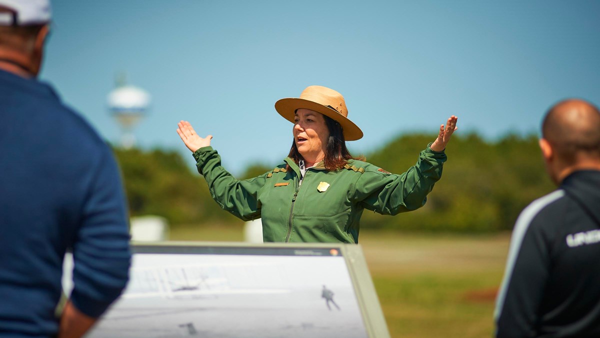A uniformed park ranger speaks in front of visitors with arms out open. 