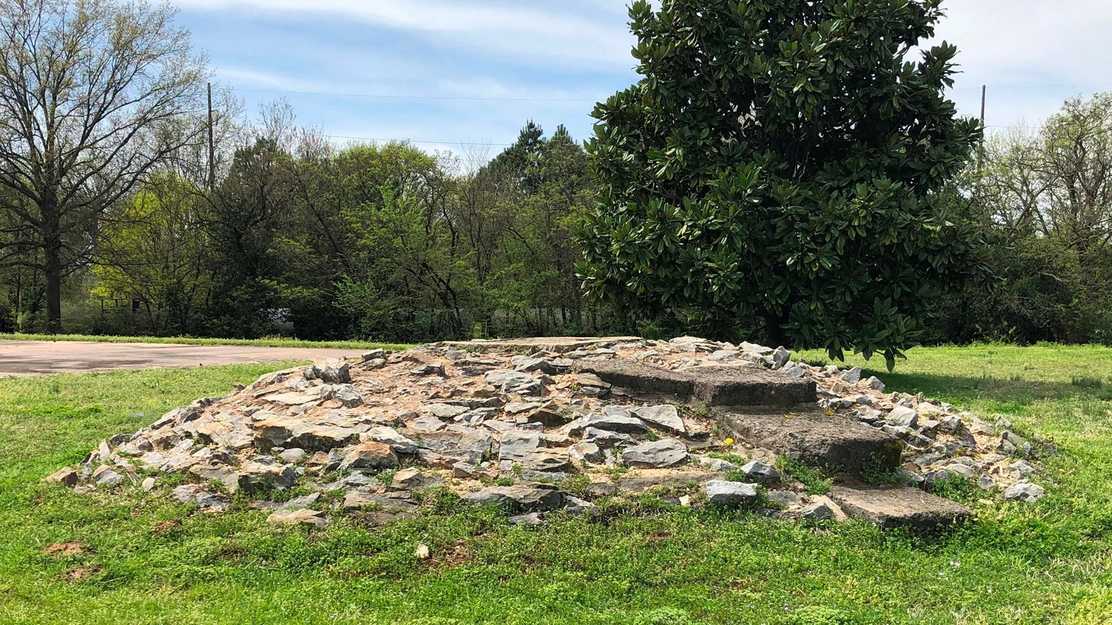 A stone mound with three small stairs surrounded by green grass with trees in the background.