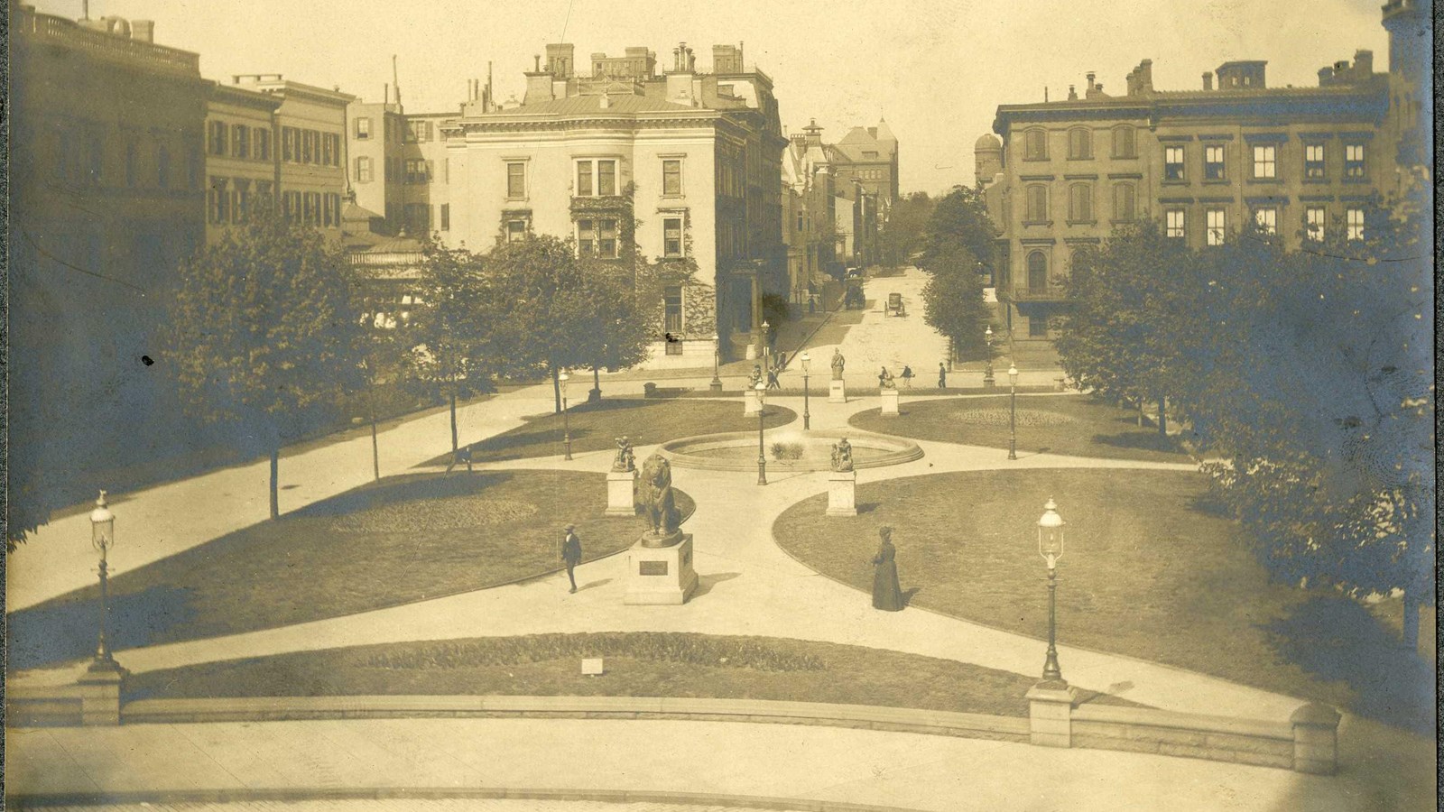 Black and white of rectangular park with fountain in middle, four half circle of grass and people 