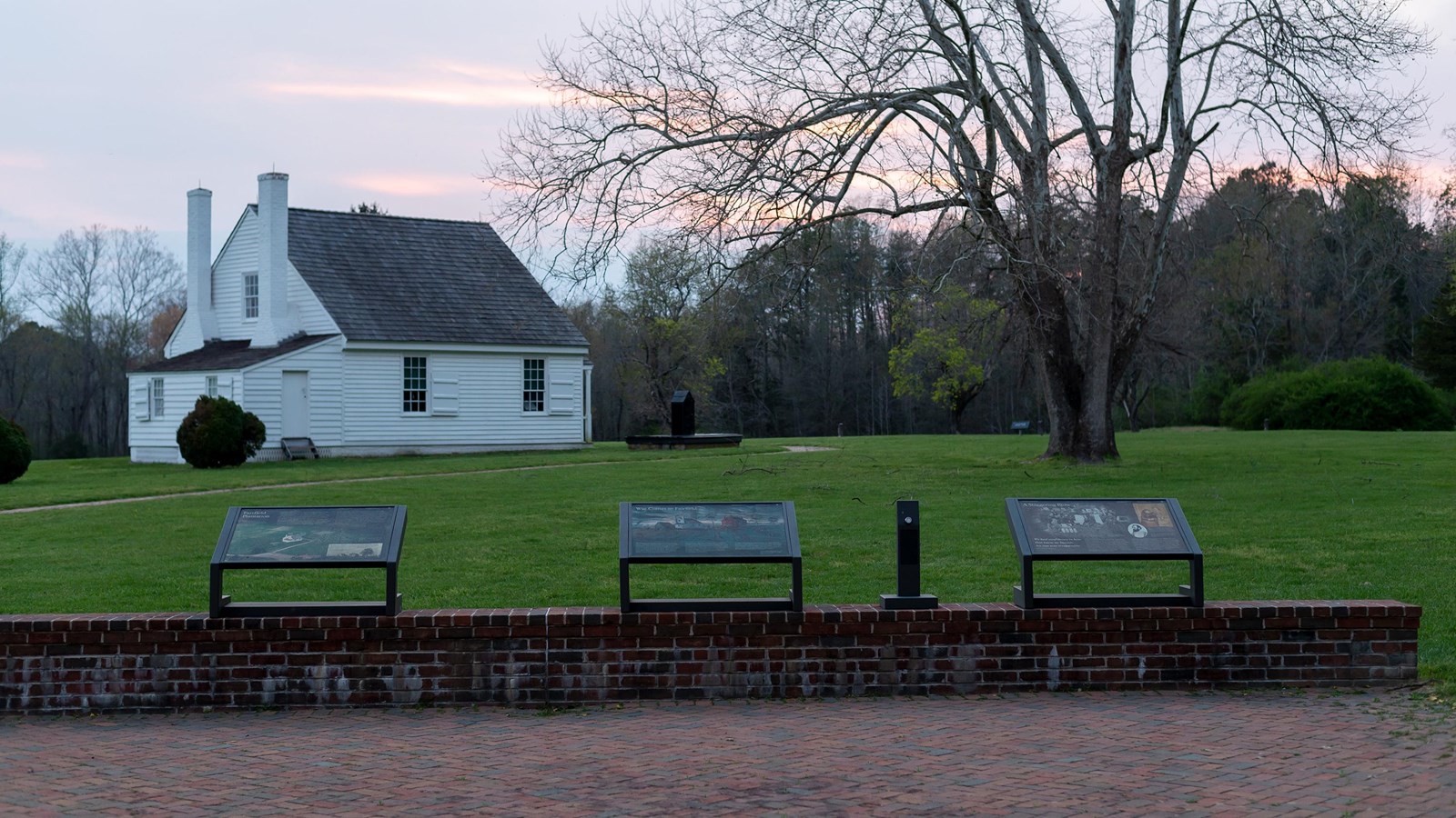Three interpretive signs in front of a brick wall looking out to a field with a white house.