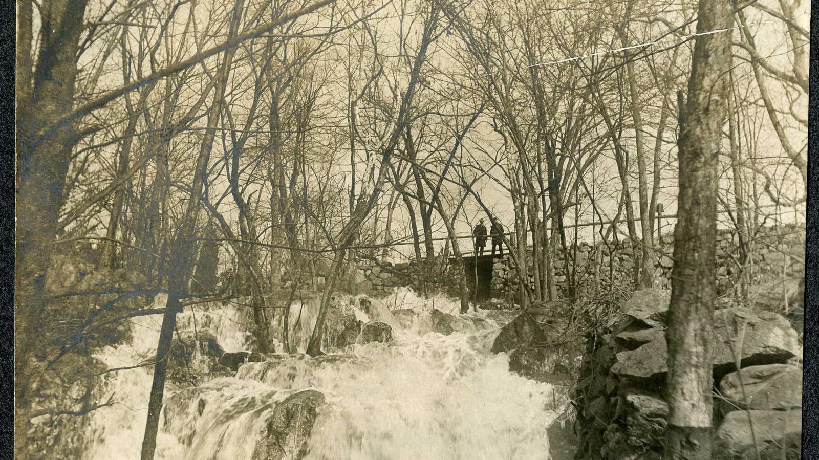 Black and white of water flowing down rocks with trees on side and people on top of water. 