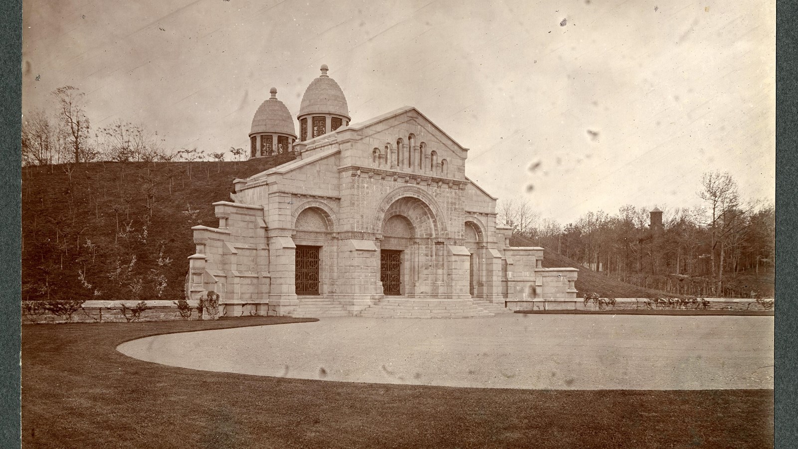 Black a white of symmetrical stone building built into hill with flat grassy area in front 