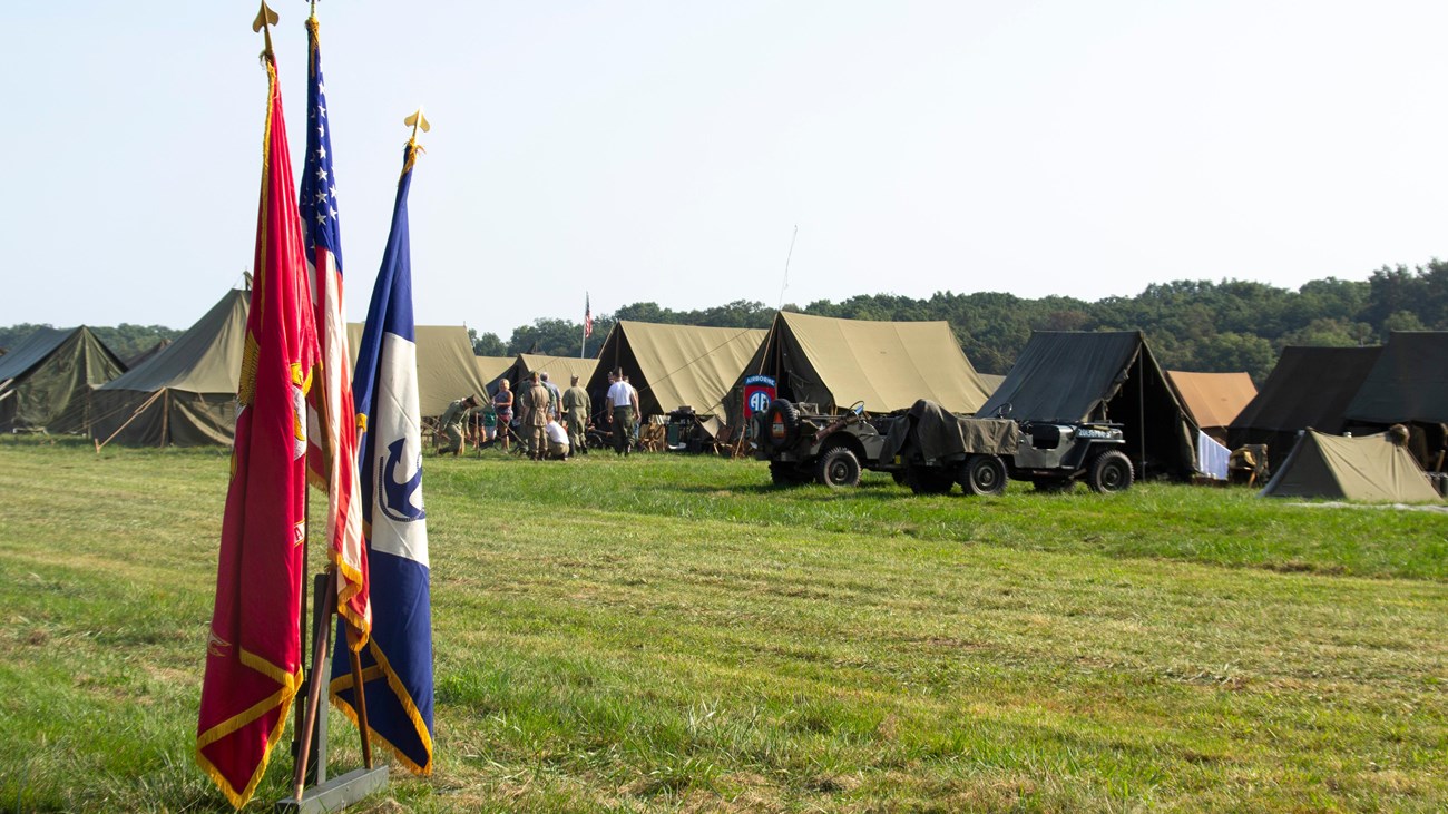 Red and blue flags stand in front of a green grass field with green army tents in the background.