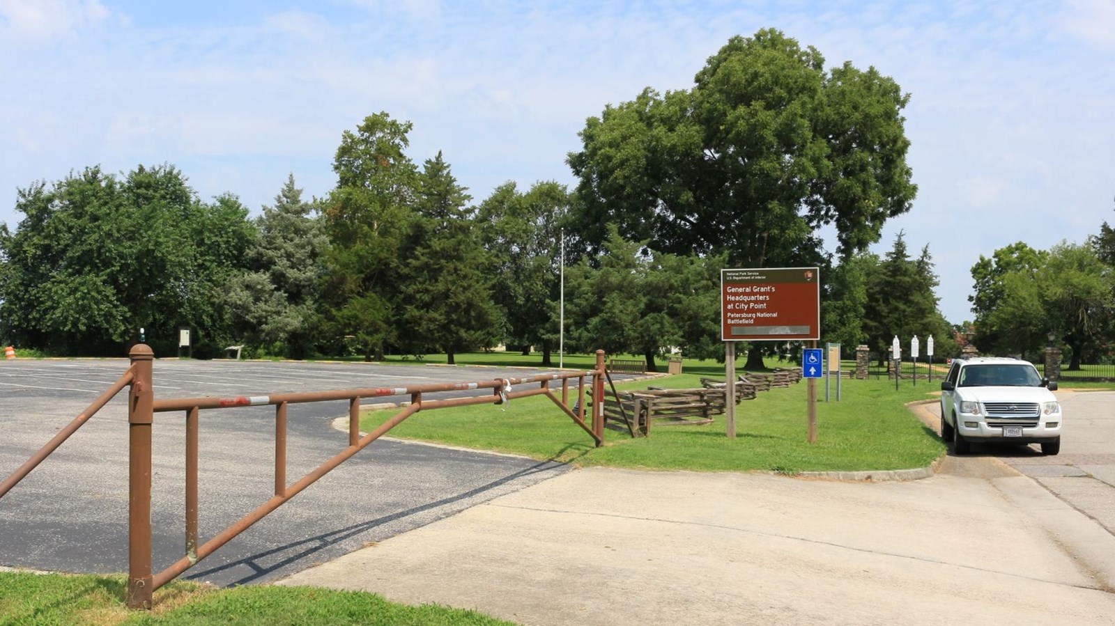 Large, paved parking lot, with brown metal gate, and brown entrance sign. White car parked near sign