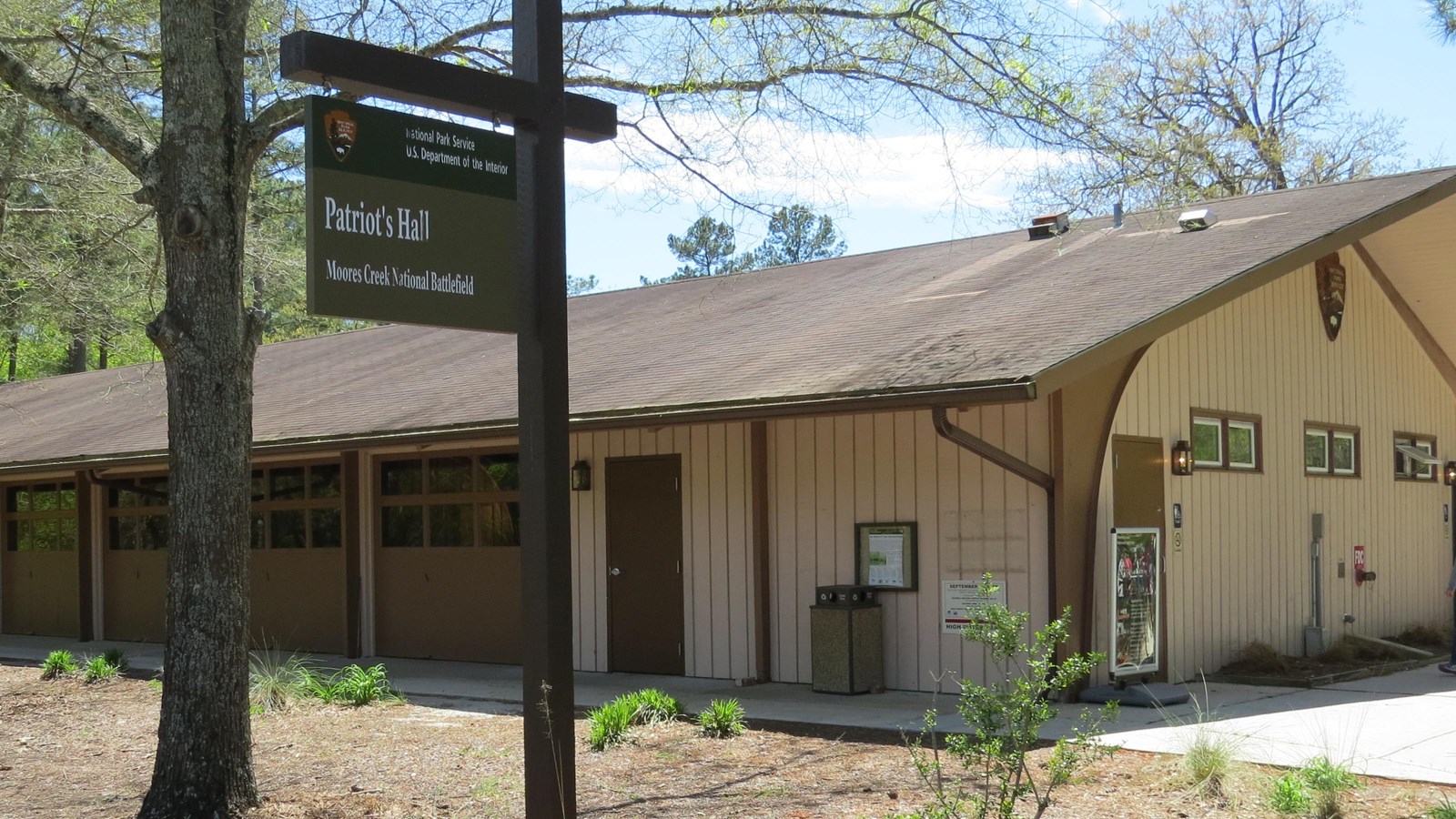 One-story building painted light brown with dirt path in the front. Wooden sign in foreground.