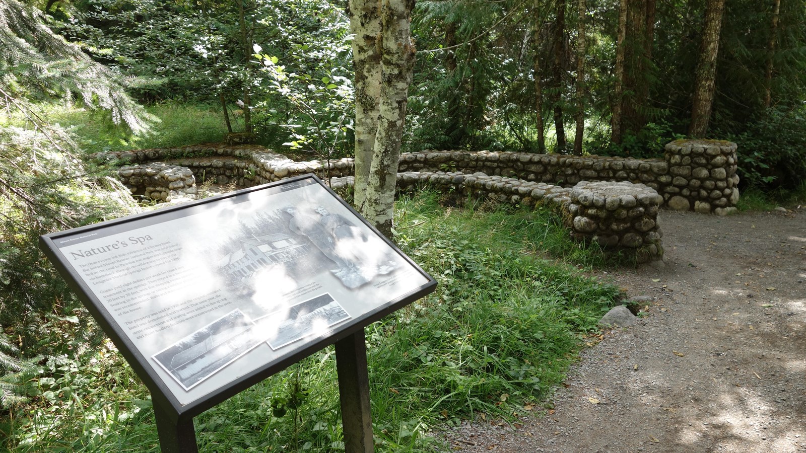 A wayside in a metal stand next to a trail bordered by a low stone wall leading to a spring.