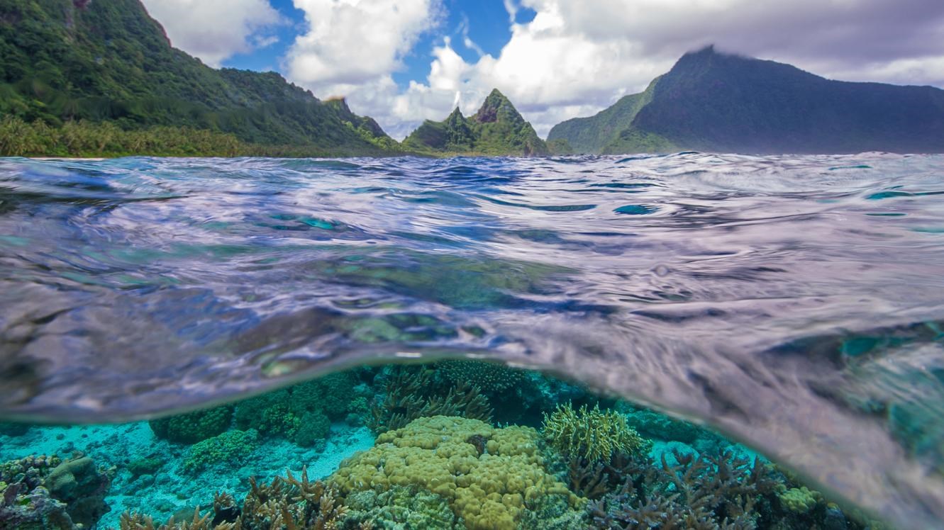 A split view above and below the surface of a mountain-ringed lagoon, with colorful coral below.