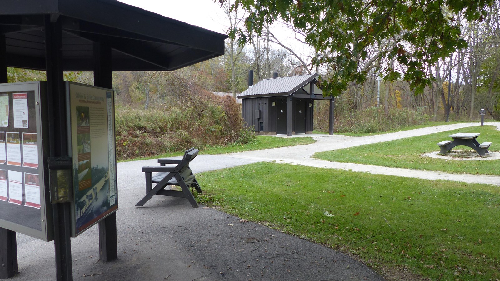 Left to right, path leads from three sided kiosk and bench, past brown restroom and picnic table.