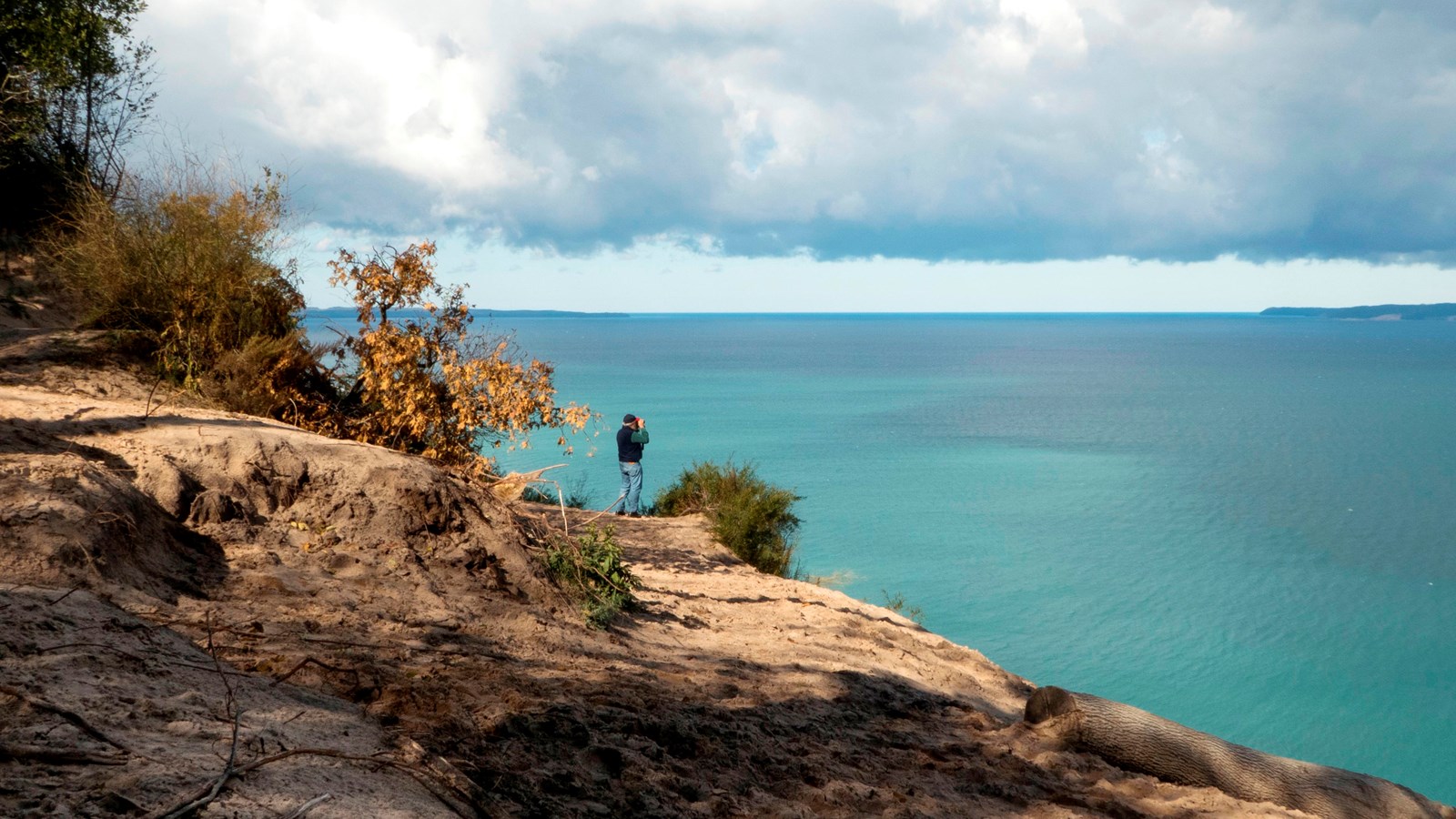 Man stands at edge of sandy dune looking out toward large body of turquoise water