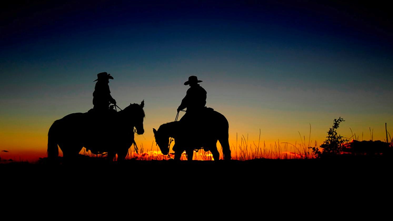 Two cowboys on horseback silhouetted against sunset.