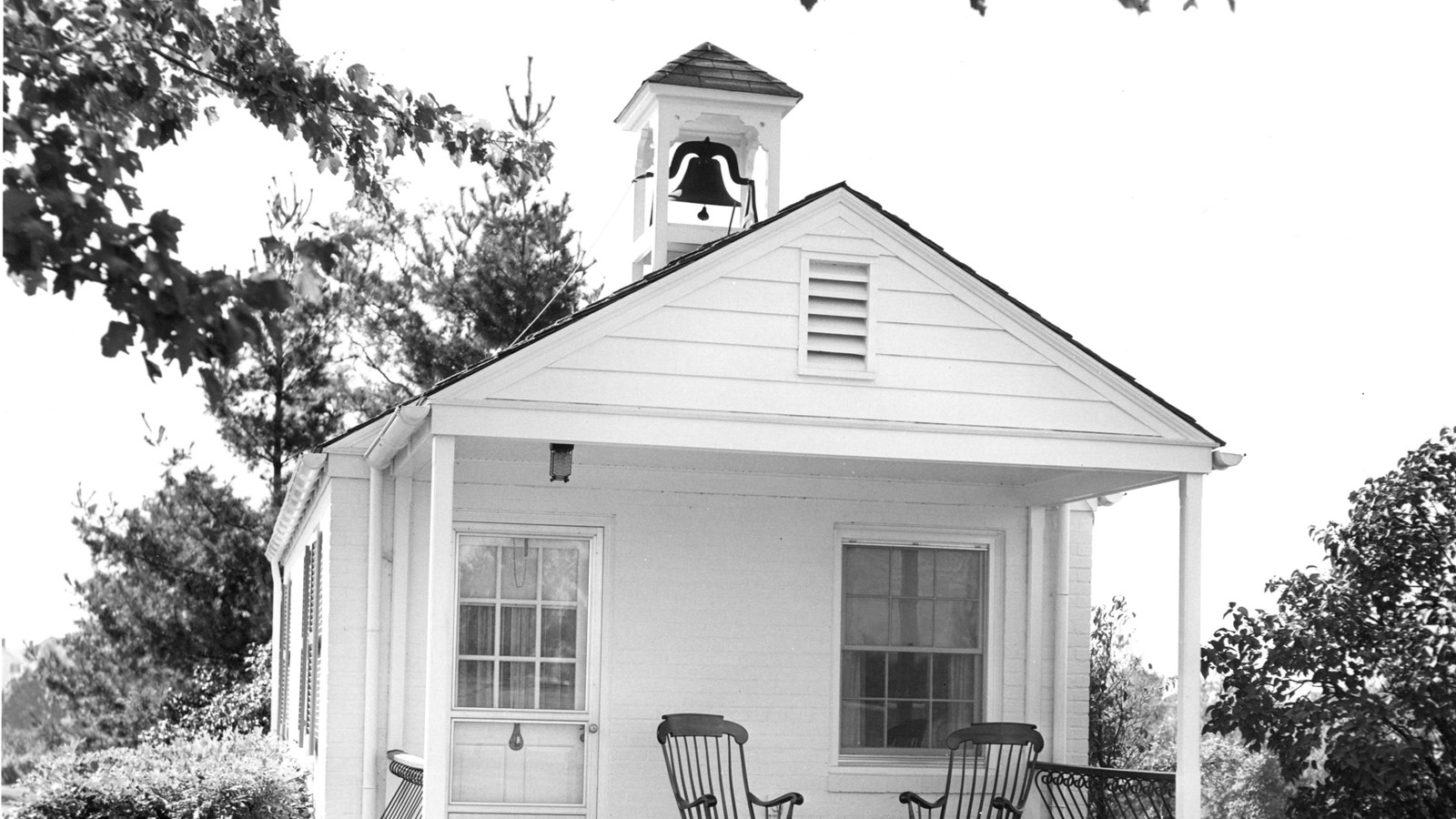 A black and white image of a white brick guesthouse with two rocking chairs in front