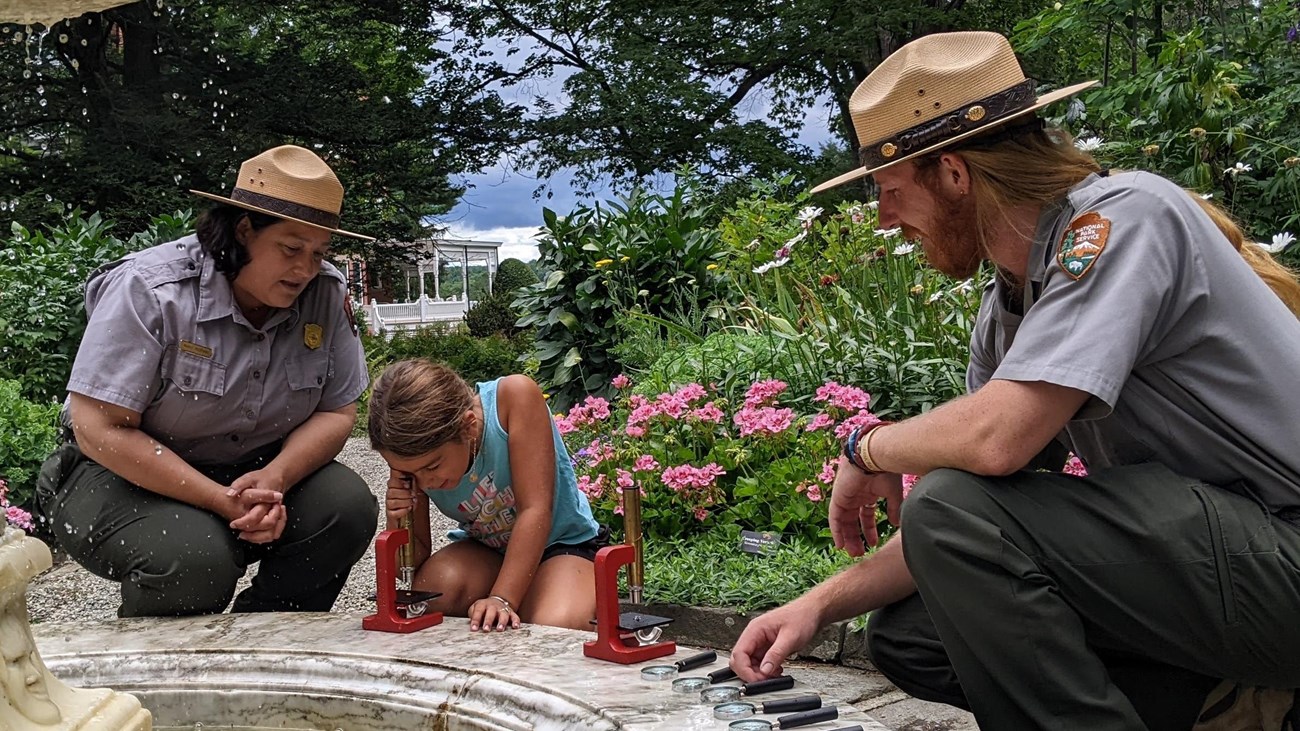 Two park rangers kneel with a child while she looks in a microscope