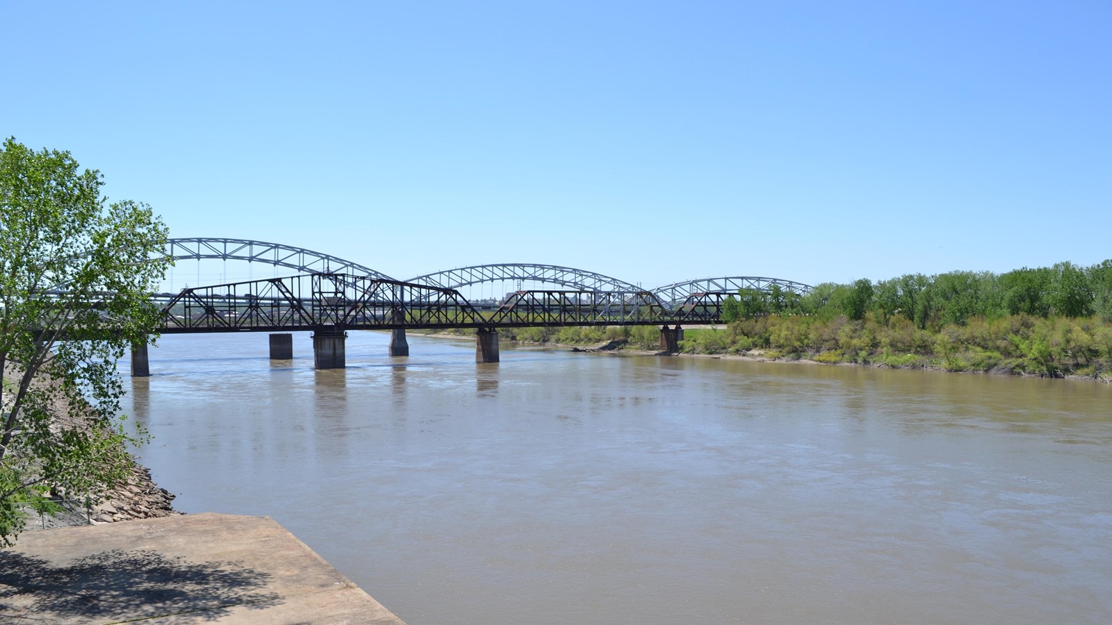 Looking out at a distant metal bridge across a flat, wide, brown river.