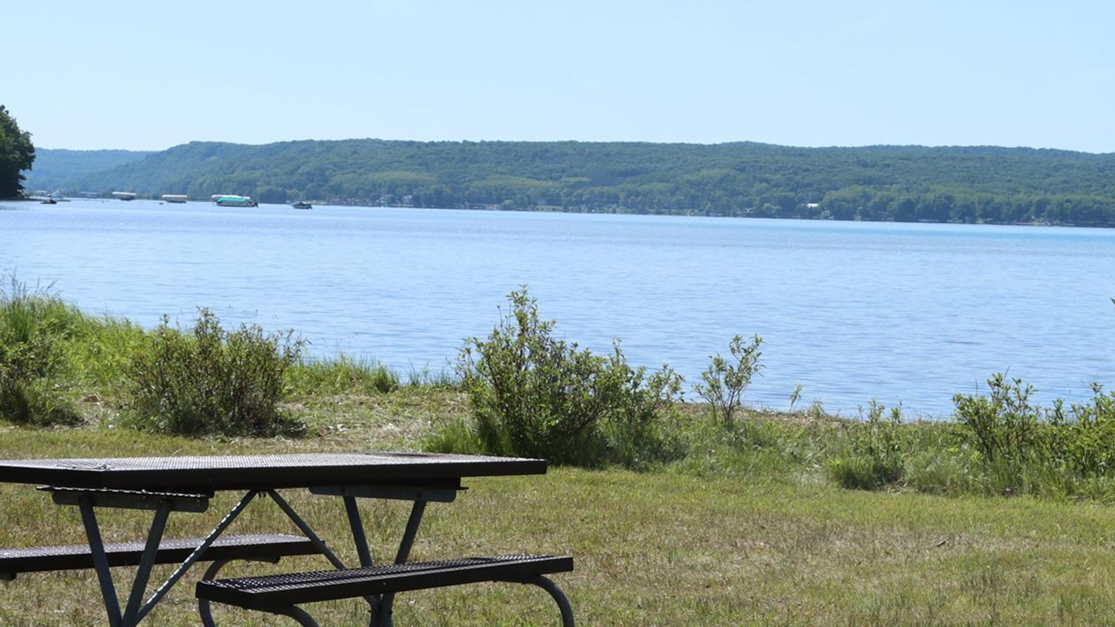 The clear blue lake opens from a grassy shoreline with a few small shrubs, picnic table and grill.