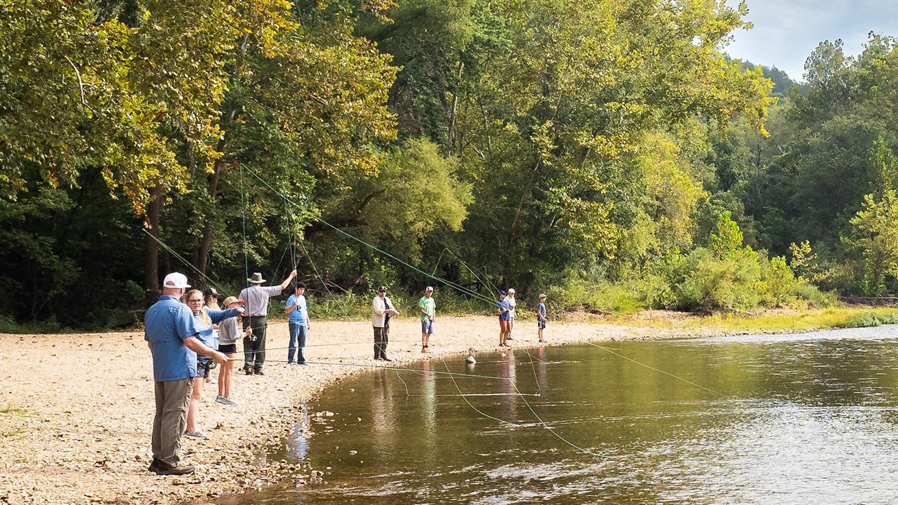 A group of visitors practice casting fly rods along the river\'s edge.