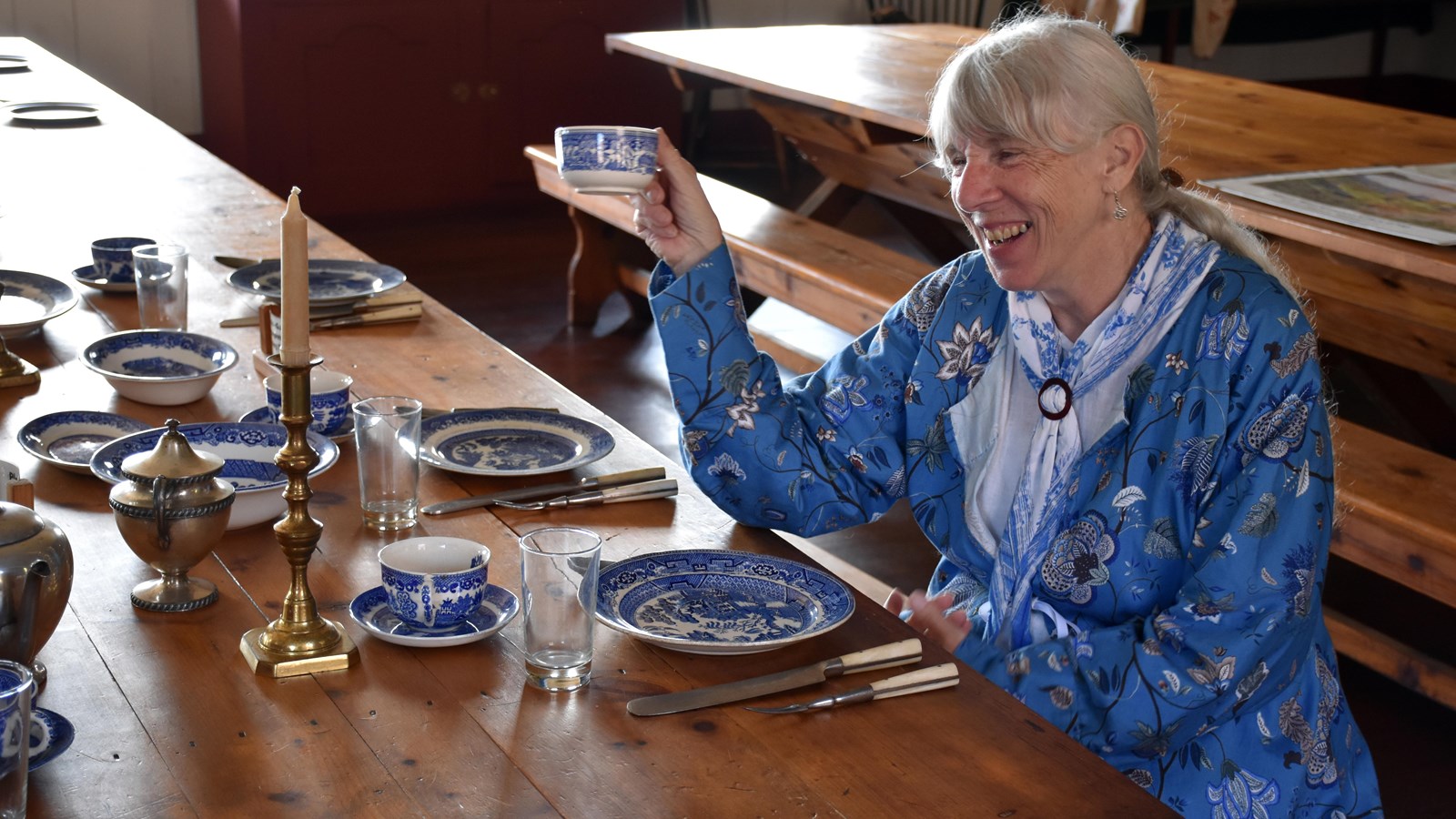 A person wearing historic clothing and holding a tea cup, sits a table with blue-patterned dishes.