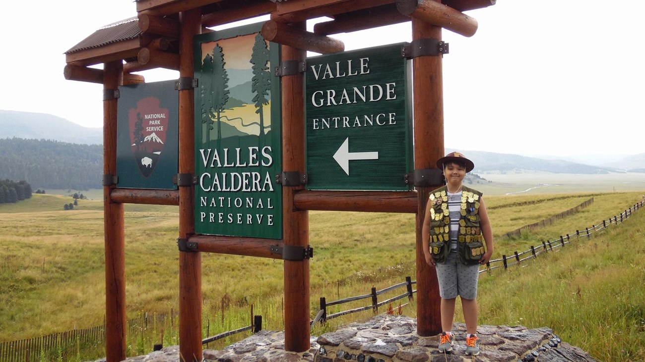 A boy wearing a Junior Ranger hat and vest filled with badges stands at Valles Caldera\'s sign.