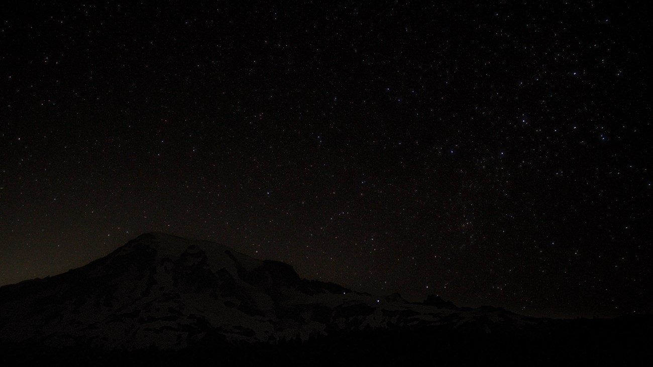 A starry sky stretches above a large volcanic peak. 