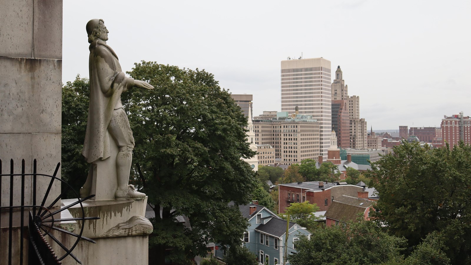 White granite statue of Roger Williams with Providence skyline in background.