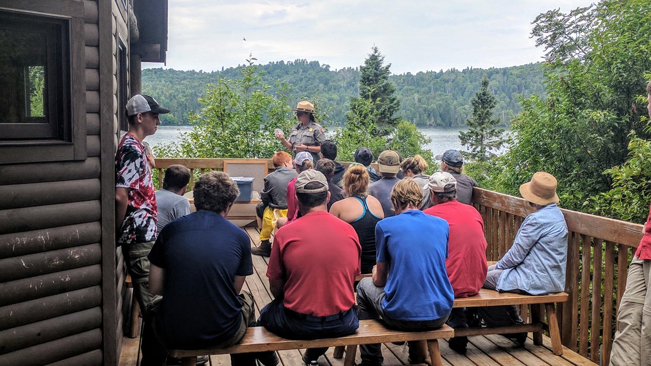 A park ranger gives a program to a group of people on a porch with Washington Harbor behind. 