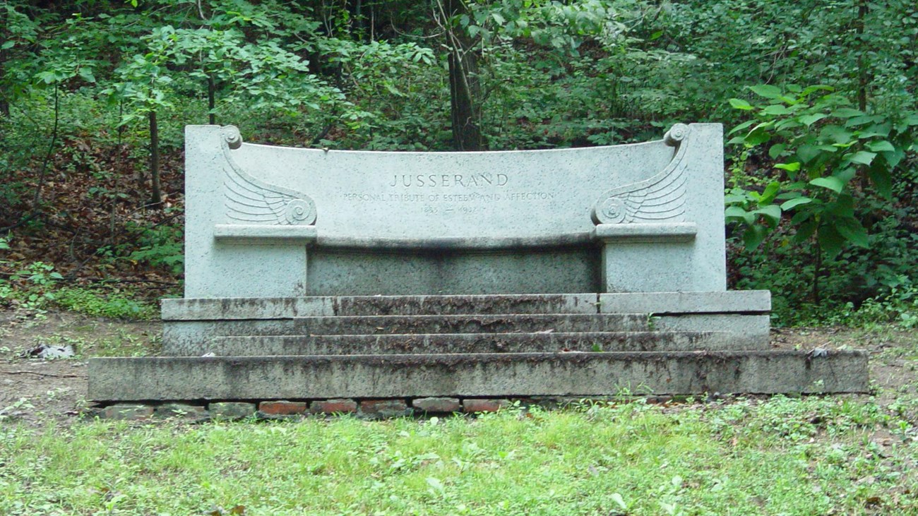 A granite bench sits atop a slight rise surrounded by green trees
