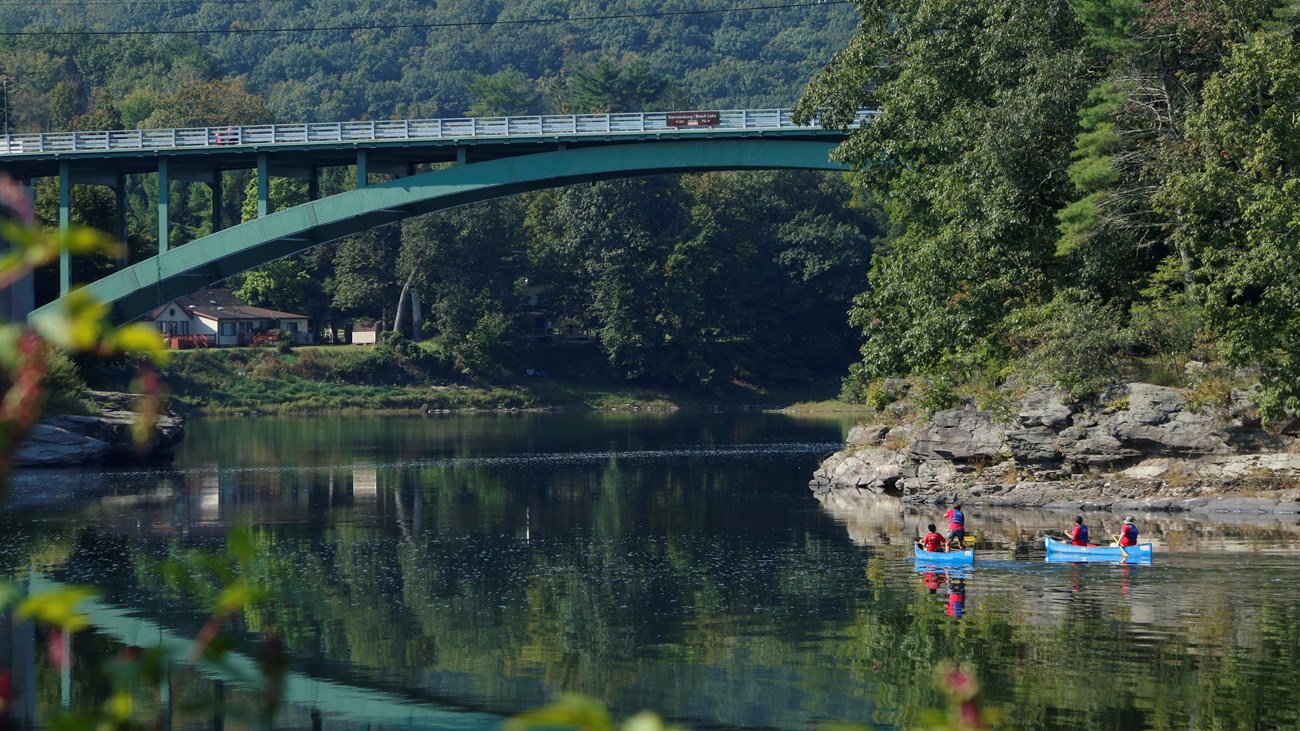 A set of 2 canoes with 2 paddlers in each canoe paddle towards the Narrowsburg Bridge.