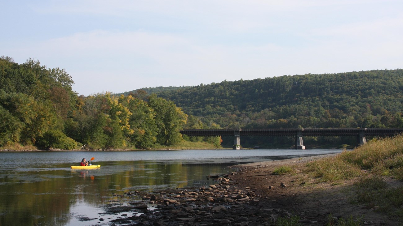 person in yellow kayak paddles in river with aqueduct-converted bridge in background.