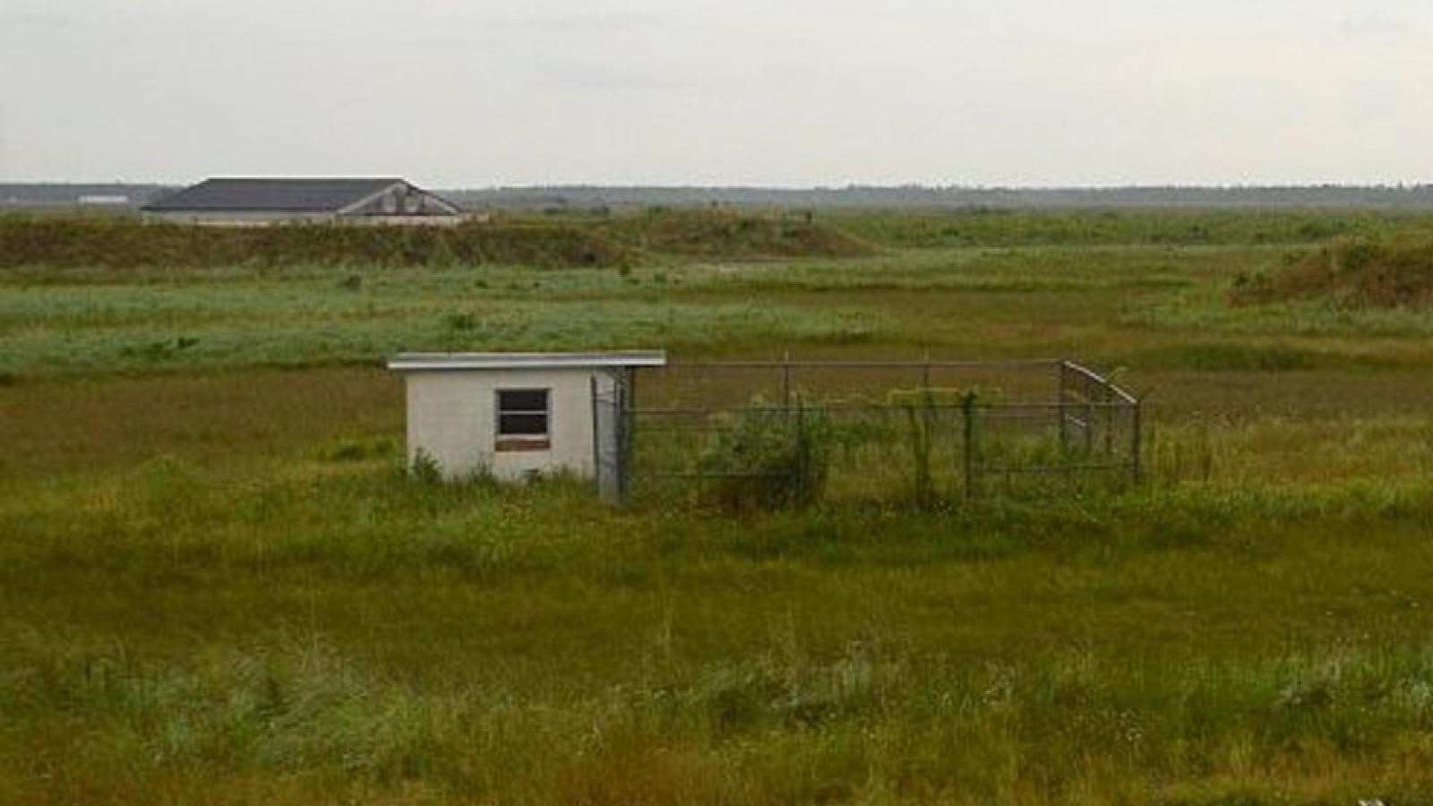 A small white building with adjacent squared in fence.  Flat landscape is covered with grasses.