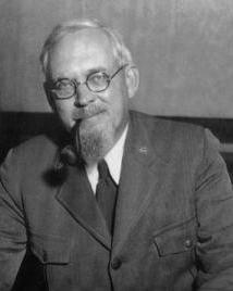 Man with beard, glasses and pipe wearing a suit poses for a picture. 