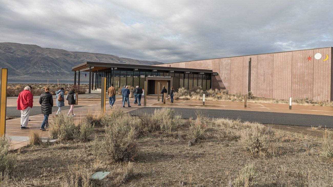 Color photograph of a building made of red stone blocks with a glassy entranceway with many people e