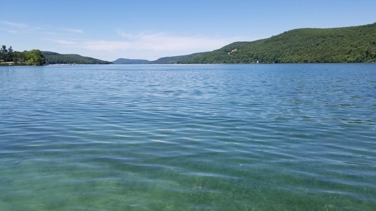 A lake with clear, blue-green water and distant tree-covered hills. 