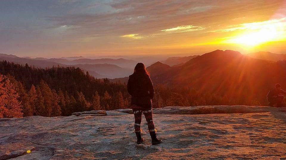 A woman stands on a granite dome watching the sunset.