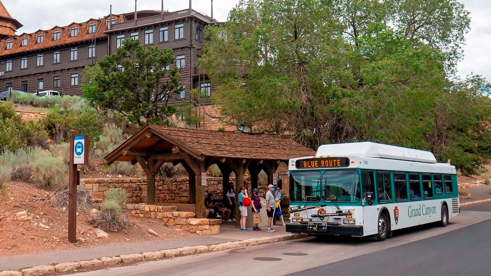 People are boarding a white bus at a shelter built into a hillside below a rustic hotel.