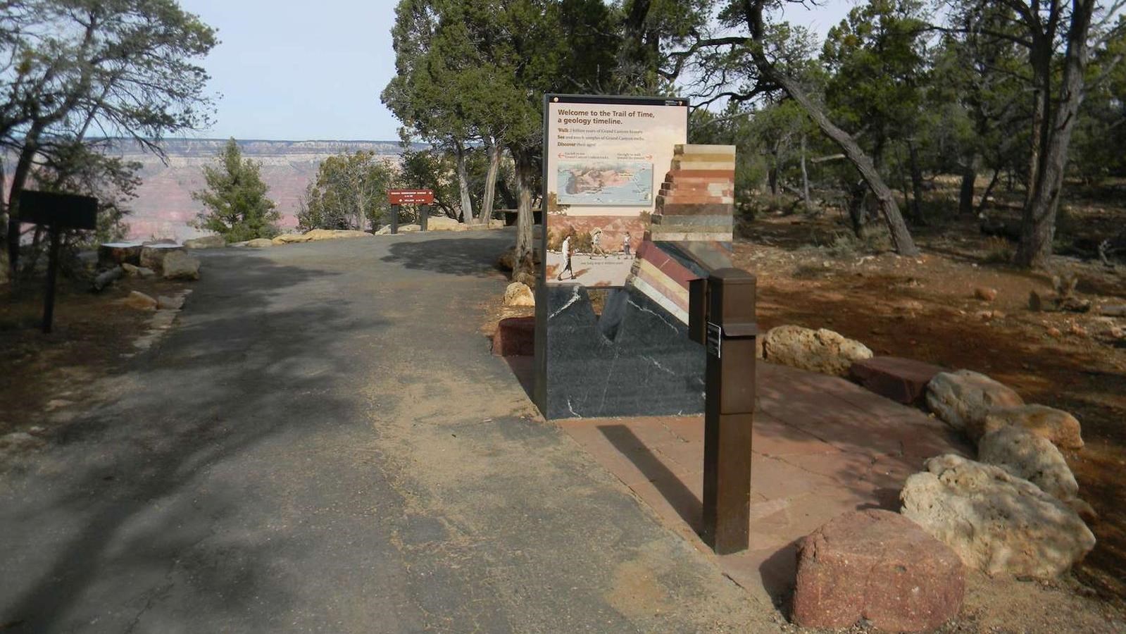 paved footpath with a stone portal sign showing canyon rock layers. Canyon landscape in the distance