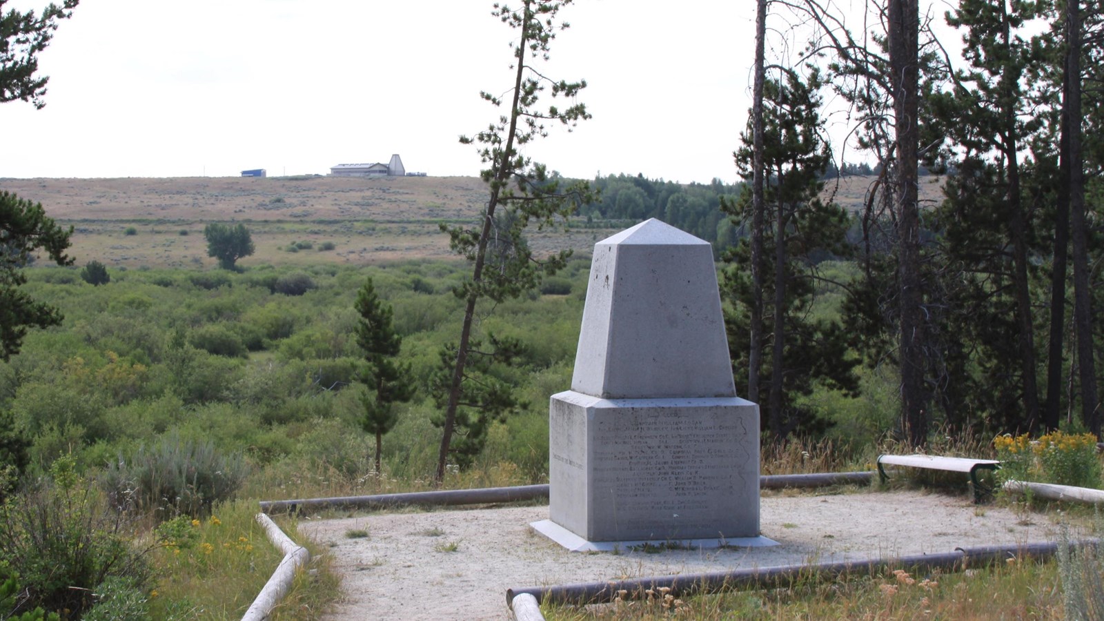 Trail leading towards 6-ton granite, obelisk shaped monument amongst lodgepole pine trees with Visit