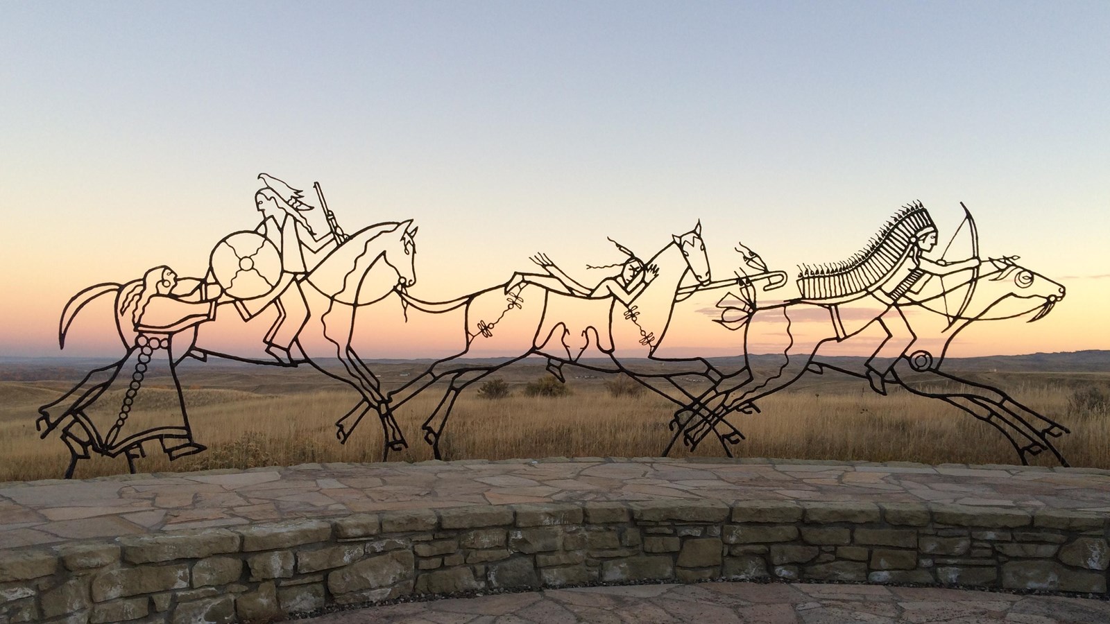A large black metal sculpture of 3 Warriors and a woman on Horseback  with sunset in background
