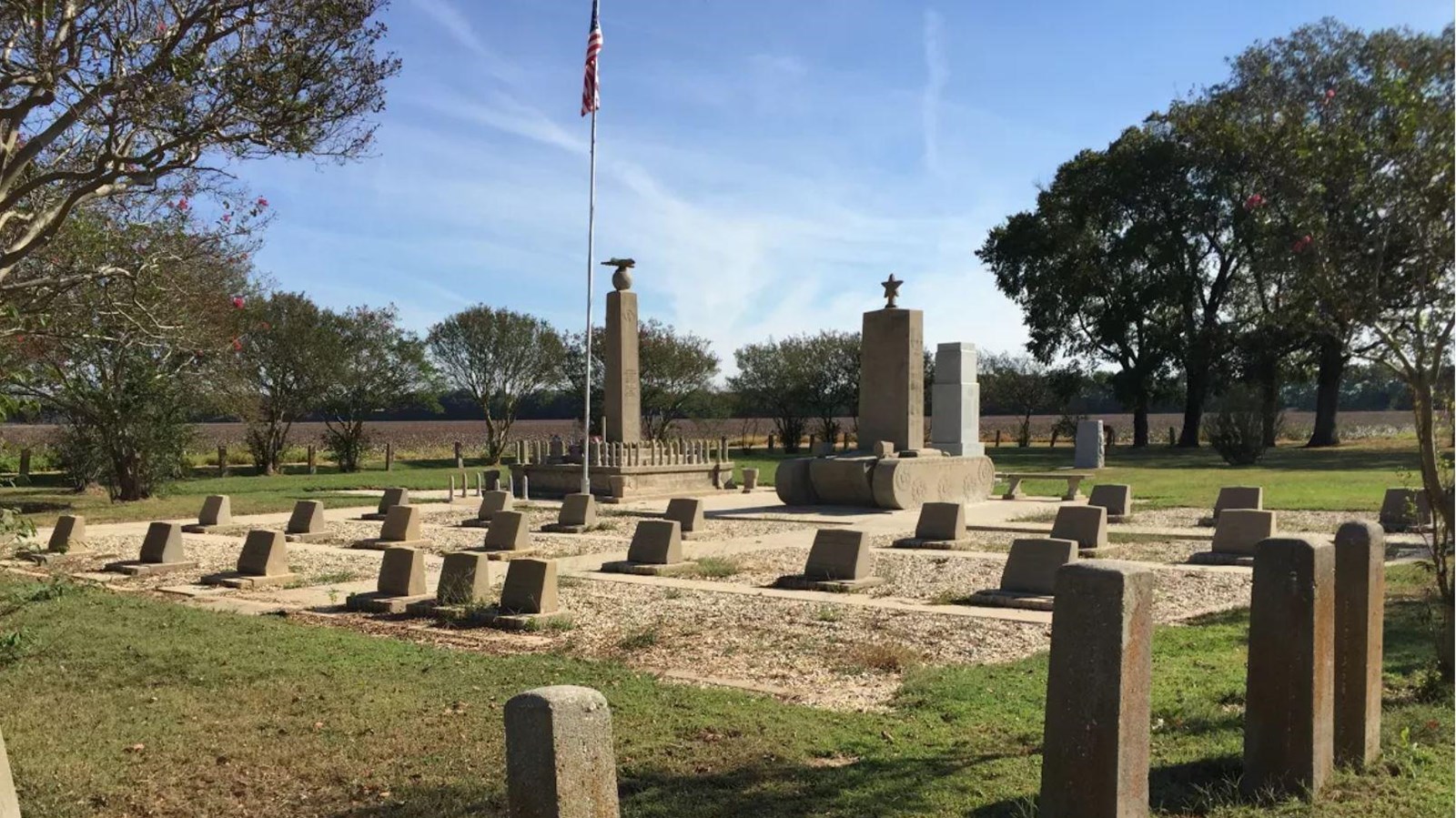 Cemetery with headstones, U.S. flag, and three monuments surrounded by grass.