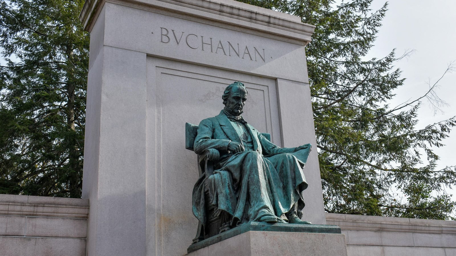 A green-tinted statue of James Buchanan on a pedestal in front of a stone wall behind it.