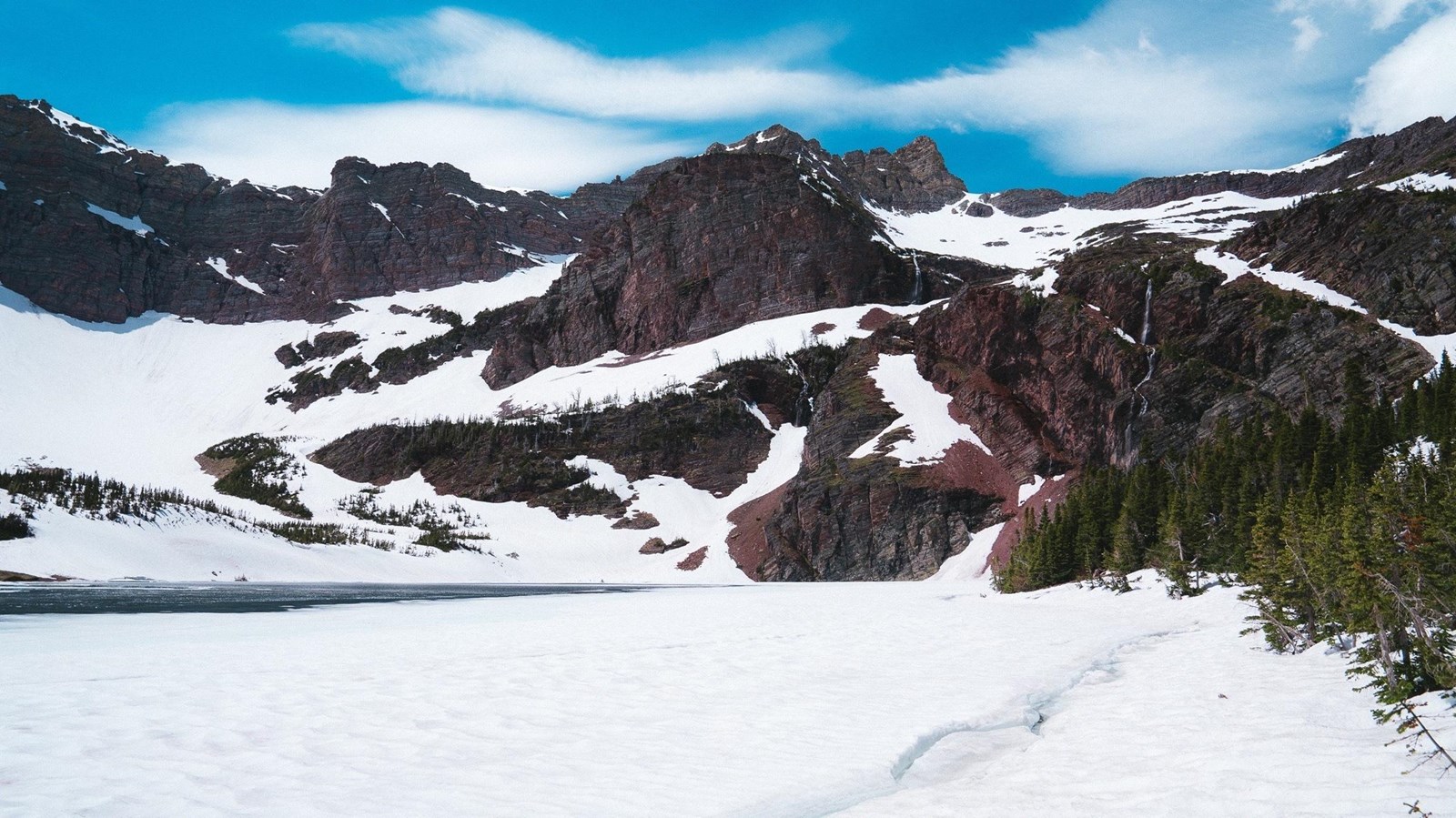 Snow covers a lake, with a small section of water visible. Red snowy cliffs are in the background.
