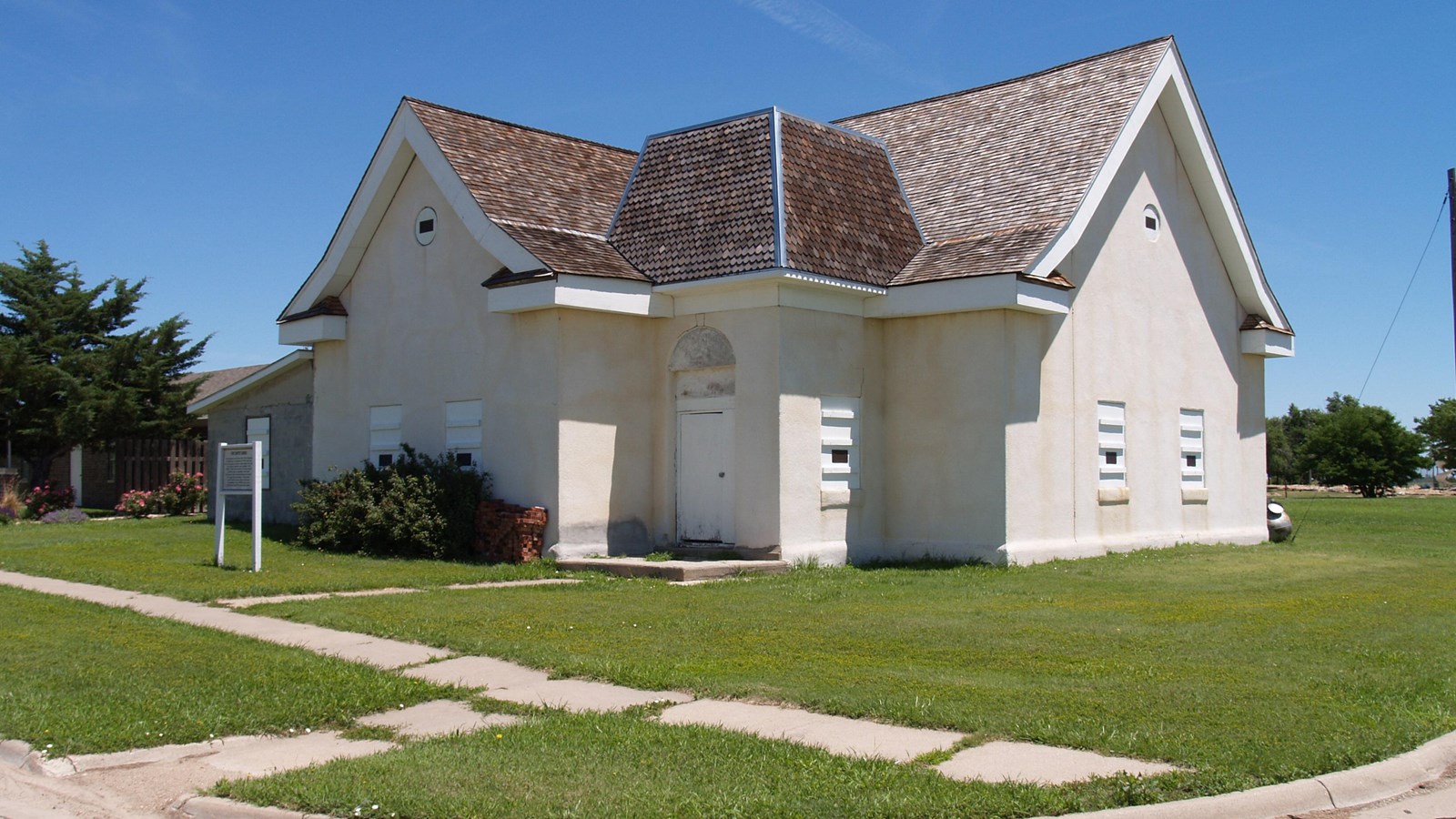 White stucco church with gray shingle roof on a grassy lawn on a street corner