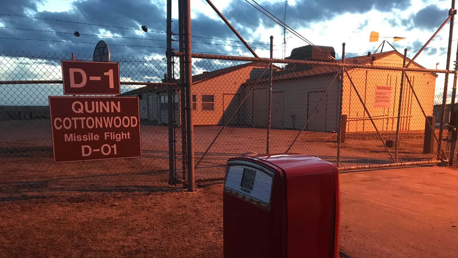a red gas pump stands outside a fenced compound with a large tan building
