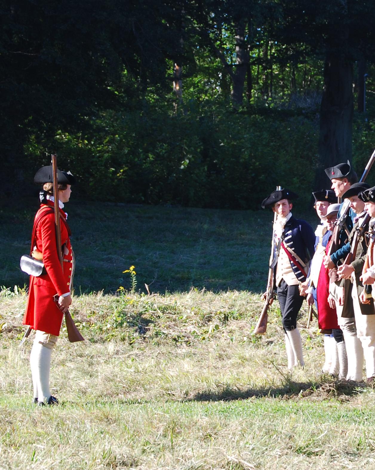 About 30 men, dressed as Revolutionary War soldiers, stand in formation in a field with muskets. 