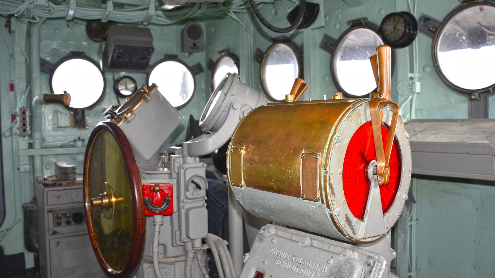 View of a green/gray painted compartment with circular windows. Brass telegraph levers in main focus