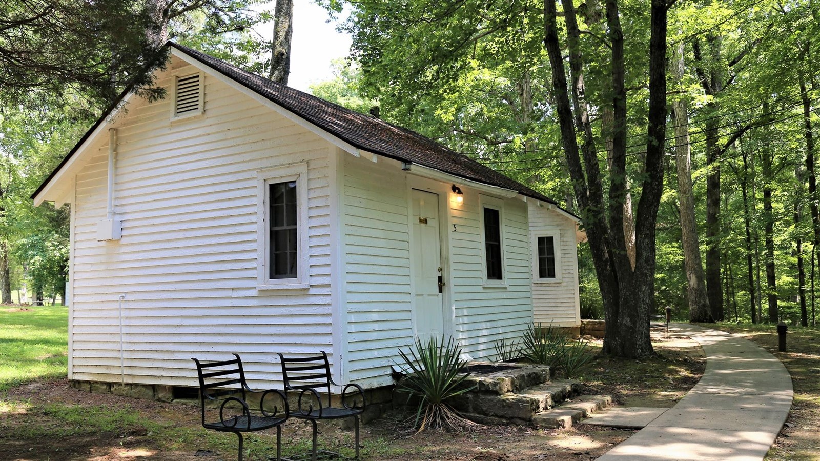 A quaint cottage with white siding framed by a green forest that shades the sidewalk.