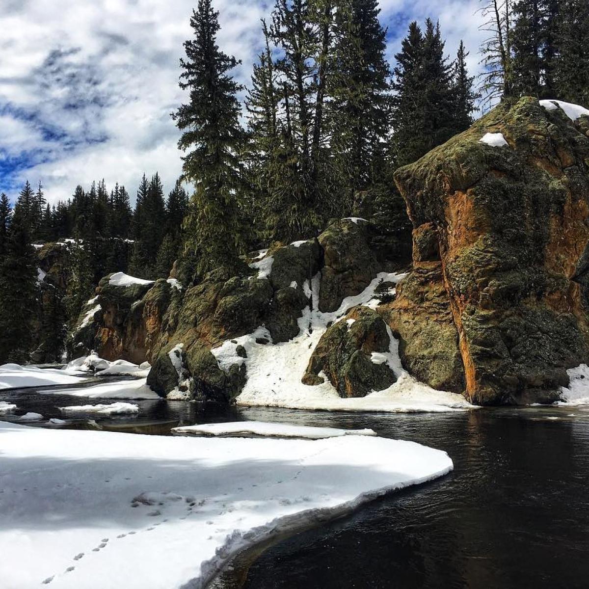 A snow-covered landscape with a small stream meandering through rock spires and evergreen trees.