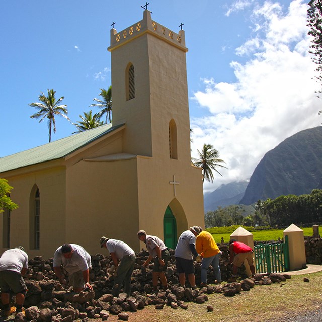 A yellow church in front of bright blue sky, people working in foreground